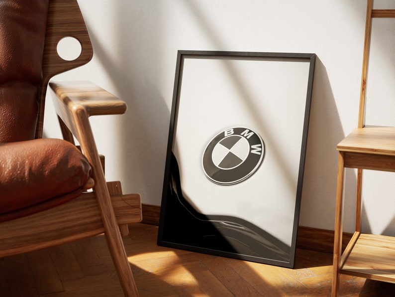 May include: Framed print featuring the BMW logo. The logo is a black and white circular emblem with the letters BMW. The print is in a black frame, leaning against a white wall. A wooden chair and side table are in the background.