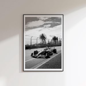 May include: A black and white framed wall art print depicting a Formula 1 race car driving on a track. The scene features palm trees in the background, a metal safety fence, and the Rolex logo visible on the trackside barriers under a cloudy sky.