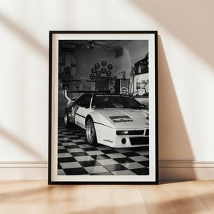 May include: Black and white framed print of a vintage race car. The car is white with black accents and sponsor logos, including "Marlboro." The image is set in a garage with a checkered floor.