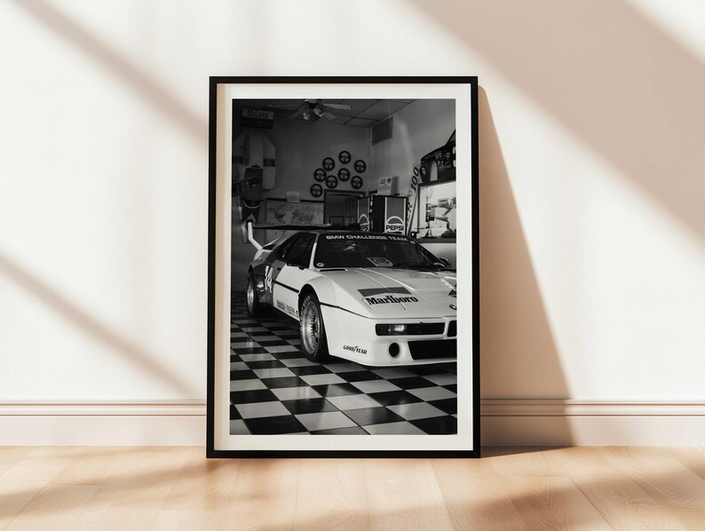 May include: Black and white framed print of a vintage race car. The car is white with black accents and sponsor logos, including "Marlboro." The image is set in a garage with a checkered floor.