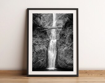 May include: Black and white framed print of a waterfall cascading down a rocky cliff. A small bridge spans the falls, surrounded by lush greenery. The print is displayed on a wooden floor.