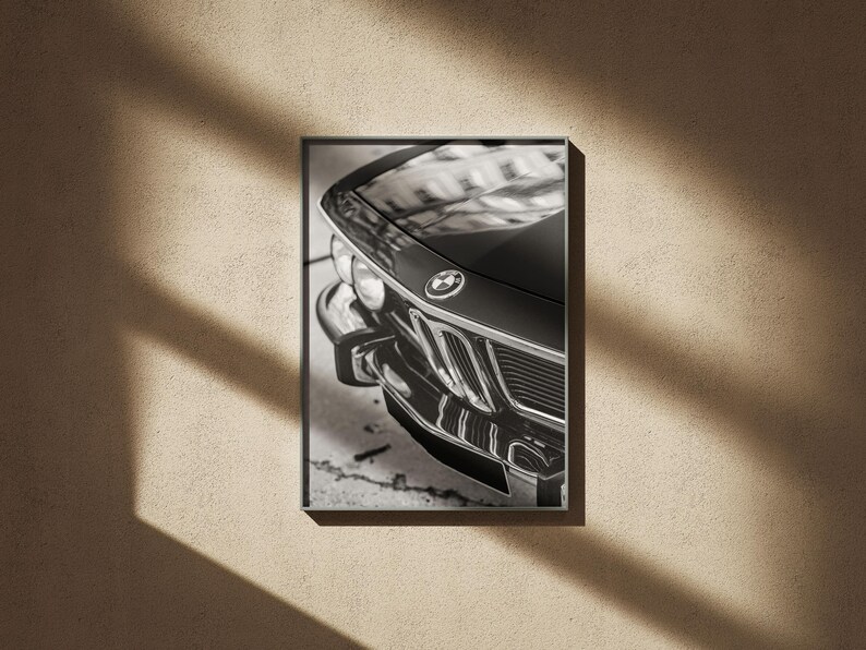 May include: Black and white framed print of a classic BMW car. The image focuses on the front of the car, showcasing the hood, headlights, and iconic BMW emblem. The car's details are highlighted by the monochrome tones.