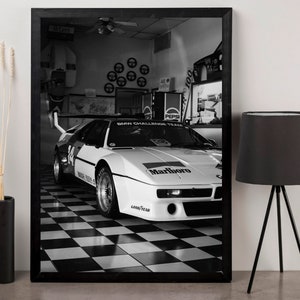 May include: Black and white framed print of a vintage race car in a garage. The car is white with black and red accents, and the Marlboro logo is visible. The checkered floor and the Pepsi machine add to the vintage feel.