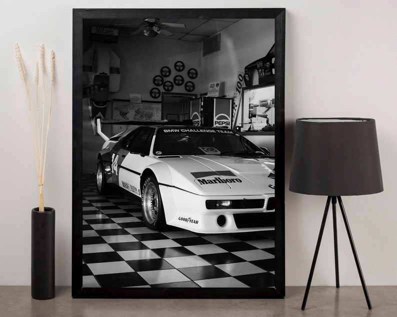 May include: Black and white framed print of a vintage race car in a garage. The car is white with black and red accents, and the Marlboro logo is visible. The checkered floor and the Pepsi machine add to the vintage feel.