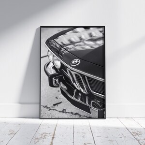 May include: Black and white framed print of a classic car's front end. The image highlights the car's chrome grille, headlights, and the BMW emblem. The car's glossy black finish reflects the surrounding environment.