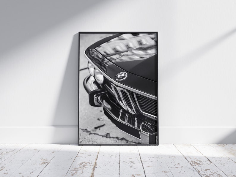 May include: Black and white framed print of a classic car's front end. The image highlights the car's chrome grille, headlights, and the BMW emblem. The car's glossy black finish reflects the surrounding environment.