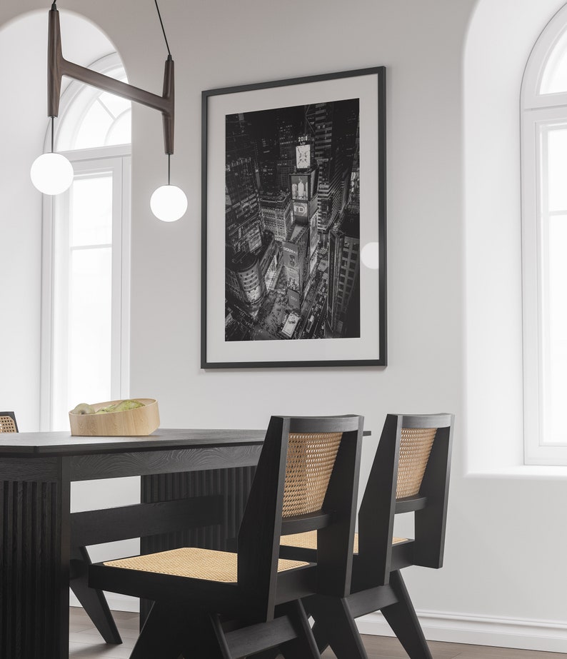 Puede incluir: Impresi&oacute;n en blanco y negro de una ciudad de noche, probablemente Nueva York, con luces brillantes y edificios altos. La impresi&oacute;n est&aacute; enmarcada en una pared blanca sobre una mesa de comedor negra con sillas negras.