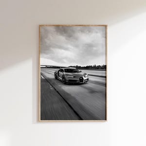 May include: Monochrome framed print featuring a silver sports car on a motorway. The car is depicted in motion, with motion blur. The sky is cloudy, creating a dynamic scene. The print is in a wooden frame.