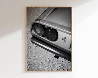 May include: Black and white framed print of a classic car's rear. The image focuses on the taillights and the car's body, with the Ferrari emblem visible. The background is a textured, gravel-like surface.