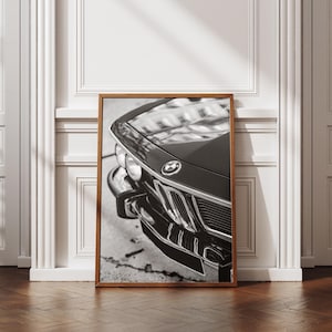 May include: Black and white framed print of a classic vintage car. The image focuses on the front of the car, showcasing the headlights, grill, and the BMW emblem. The car's hood reflects the surrounding environment.