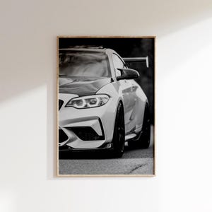 Puede incluir: Fotografía en blanco y negro de un coche deportivo blanco con detalles en negro. El coche se muestra de frente, destacando sus faros y diseño aerodinámico. La imagen está enmarcada.