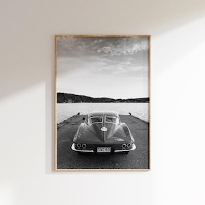 Pode incluir: Impressão emoldurada em preto e branco de um carro clássico vintage estacionado em um cais, de costas para o espectador. O carro tem um design elegante com faróis visíveis e a placa diz "SWC-63". O fundo apresenta um lago e um céu nublado.
