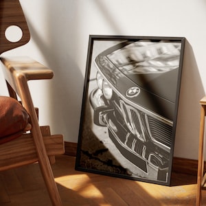 May include: Black and white framed print of a classic BMW car. The image focuses on the front of the vehicle, showcasing the grille, headlights, and emblem. The print is displayed against a white wall, next to a wooden chair.