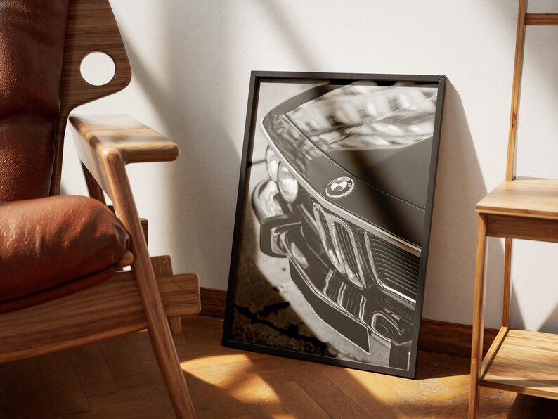 May include: Black and white framed print of a classic BMW car. The image focuses on the front of the vehicle, showcasing the grille, headlights, and emblem. The print is displayed against a white wall, next to a wooden chair.