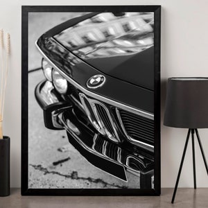 May include: Black and white framed print of a classic car's front end. The image highlights the car's hood, grille, and headlights, with the BMW logo prominently displayed. The glossy black paint reflects the surrounding environment.