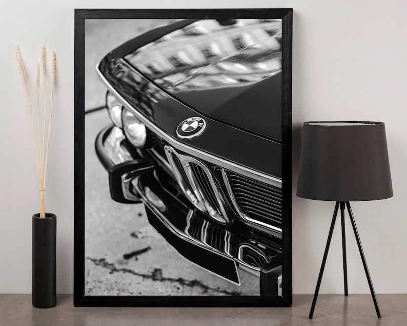 May include: Black and white framed print of a classic car's front end. The image highlights the car's hood, grille, and headlights, with the BMW logo prominently displayed. The glossy black paint reflects the surrounding environment.