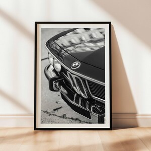 May include: Black and white framed print of a classic car's front end. The image highlights the car's hood, grill, and bumper, showcasing its vintage design. The BMW emblem is visible on the hood.