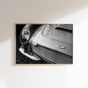 May include: Black and white framed print of a classic car's front end. The image highlights the car's headlight, grille, and hood, showcasing its sleek design. The car's emblem is visible on the hood.