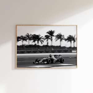 Puede incluir: Impresión enmarcada en blanco y negro de un coche de Fórmula 1 corriendo en una pista. Se ven palmeras borrosas en el fondo. La imagen está en un marco de madera y montada en una pared blanca.