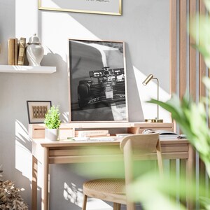 May include: A light wooden desk with a framed monochrome photograph of a racing car. A small potted plant, books, and a gold desk lamp are on the desk. A white shelf holds books and a decorative vase.