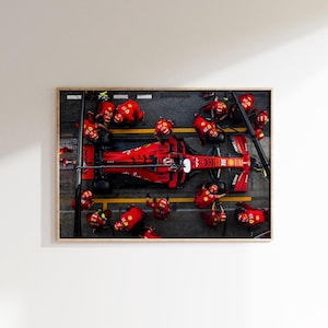 May include: A framed print of a Formula 1 race car in a pit stop. The red car is surrounded by crew members in red uniforms and helmets. The image is taken from an overhead perspective.