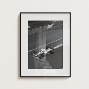 May include: Black and white framed print of a Formula 1 race car speeding on a track. The car is white with black accents, and a trail of smoke follows it. The image is a high-angle shot.