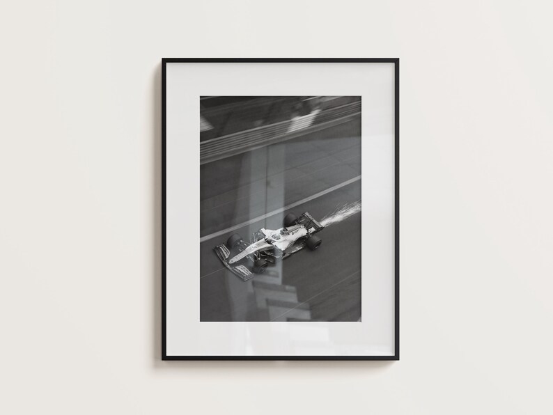 May include: Black and white framed print of a Formula 1 race car speeding on a track. The car is white with black accents, and a trail of smoke follows it. The image is a high-angle shot.