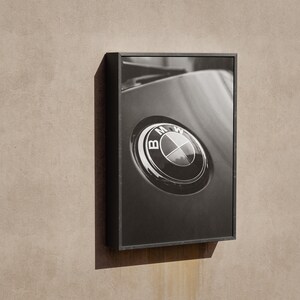 Puede incluir: Fotograf&iacute;a en blanco y negro enmarcada de un emblema de coche BMW. El emblema es circular con el logotipo de BMW en el centro. La fotograf&iacute;a est&aacute; en un marco negro y montada en una pared beige.
