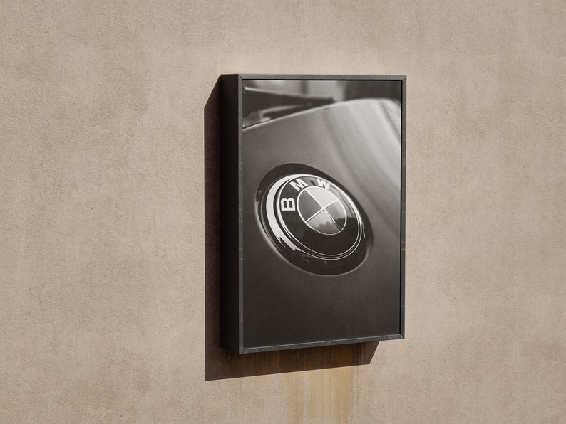 Puede incluir: Fotograf&iacute;a en blanco y negro enmarcada de un emblema de coche BMW. El emblema es circular con el logotipo de BMW en el centro. La fotograf&iacute;a est&aacute; en un marco negro y montada en una pared beige.