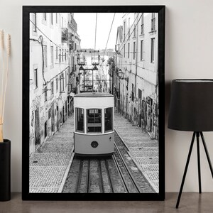 May include: Black and white framed print of a vintage streetcar traveling down a cobblestone street. The streetcar is light yellow and white, with a detailed view of the buildings on either side. The image is framed in a black border.