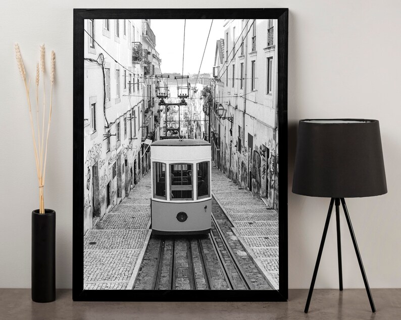 May include: Black and white framed print of a vintage streetcar traveling down a cobblestone street. The streetcar is light yellow and white, with a detailed view of the buildings on either side. The image is framed in a black border.
