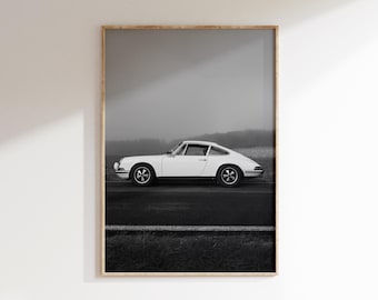 May include: Framed black and white print of a classic white sports car on a road. The car is a coupe with a sleek design, set against a foggy backdrop. The print is in a wooden frame.