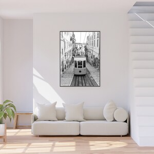 May include: Black and white framed print of a cable car traveling down a cobblestone street, flanked by buildings. The image is a vertical composition, with the cable car centered on the tracks.