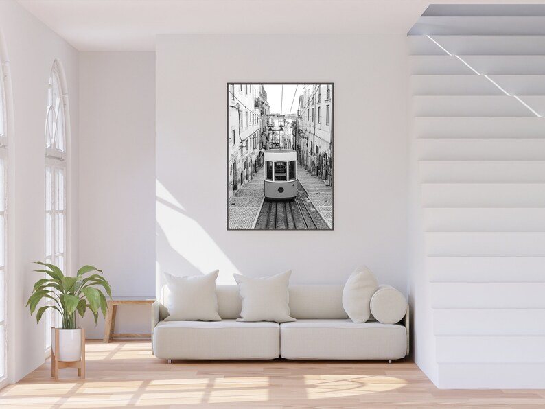 May include: Black and white framed print of a cable car traveling down a cobblestone street, flanked by buildings. The image is a vertical composition, with the cable car centered on the tracks.