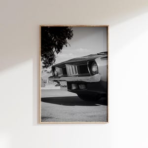May include: Framed monochrome print of a classic car's front. The car features a distinctive grille, headlights, and bumper. The number plate reads "cuda". The image is set against a backdrop of a tree and a cloudy sky.