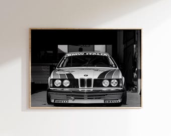 May include: A black and white photograph of a BMW race car parked in a garage. The front view shows the iconic kidney grille, round headlights, and a windscreen banner reading BMW ITALIA. Pirelli branding is visible on the front bumper.