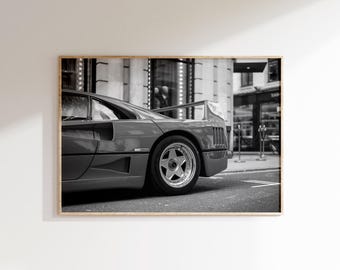 May include: Black and white framed print of a classic sports car. The car is dark gray with a prominent rear spoiler and five-spoke wheels. The image is set against a city backdrop, with buildings and a street visible.