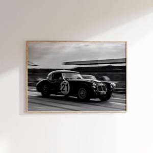 May include: Black and white framed print of a vintage race car speeding on a track. The car has the number 21 on its side and is in motion, creating a sense of speed. The image is a classic, capturing the essence of motorsport.