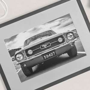 Pode incluir: Impress&atilde;o emoldurada a preto e branco de um Ford Mustang cl&aacute;ssico. O carro est&aacute; centrado, mostrando a frente com far&oacute;is, grelha e placa de matr&iacute;cula. A moldura &eacute; cinzenta escura e a imagem est&aacute; sobre um passe-partout cinzento claro.