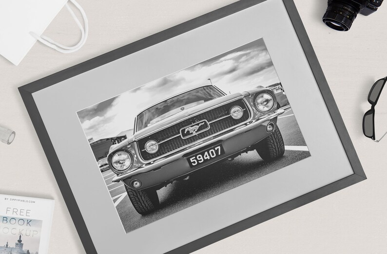 Pode incluir: Impress&atilde;o emoldurada a preto e branco de um Ford Mustang cl&aacute;ssico. O carro est&aacute; centrado, mostrando a frente com far&oacute;is, grelha e placa de matr&iacute;cula. A moldura &eacute; cinzenta escura e a imagem est&aacute; sobre um passe-partout cinzento claro.