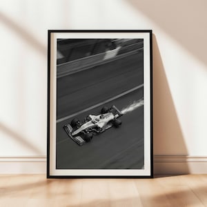 May include: Black and white framed print of a Formula 1 race car speeding along a track. The car is in motion, with a blurred background and a trail of vapor. The print is displayed on a wooden floor against a white wall.