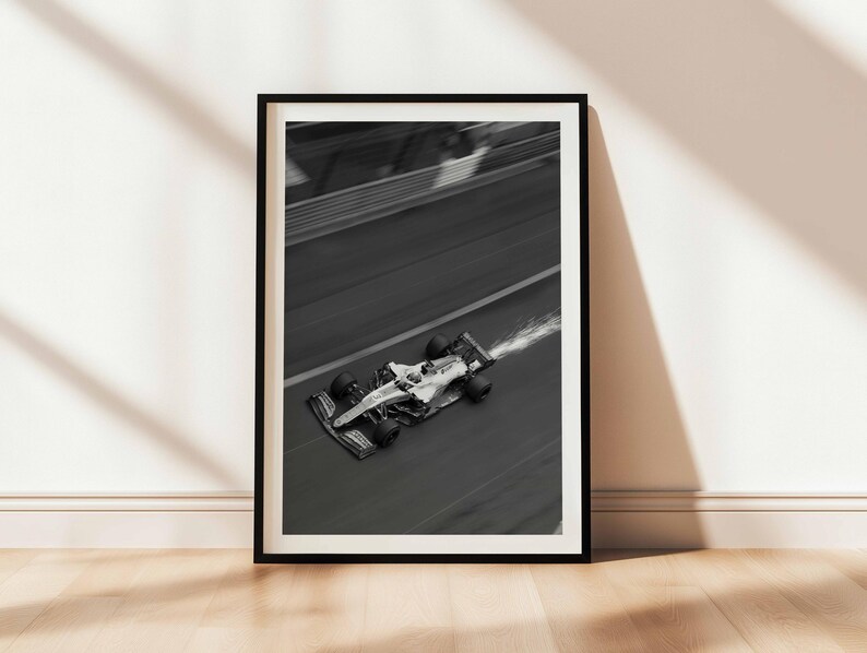 May include: Black and white framed print of a Formula 1 race car speeding along a track. The car is in motion, with a blurred background and a trail of vapor. The print is displayed on a wooden floor against a white wall.