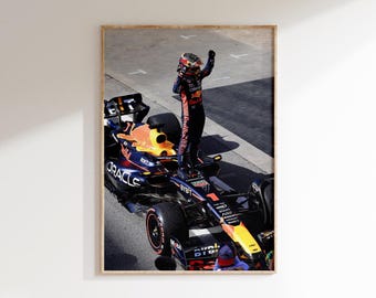 May include: Framed print of a Formula 1 race car with a driver standing on top, raising their arm in celebration. The car is primarily navy blue, orange, and yellow, with sponsor logos. The driver wears a helmet and racing suit.