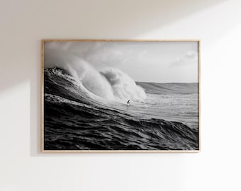 May include: Black and white framed print of a surfer riding a large wave. The wave is breaking, creating a wall of water. The surfer is a small silhouette against the wave. The ocean is dark and choppy.