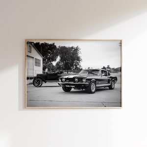 May include: Black and white photograph of two classic cars. A black Shelby Mustang with white racing stripes is parked next to a vintage black car. The cars are on a concrete surface, with a building and trees in the background.