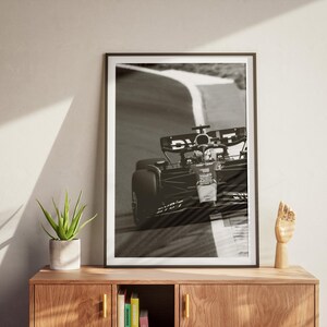 May include: Monochrome framed print of a Formula 1 car on a race track. The print is displayed on a wooden cabinet, alongside a potted plant and a wooden hand sculpture.