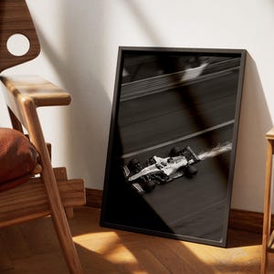 May include: Black and white framed print of a Formula 1 race car speeding on a track. The image is angled, showing the car in motion with a blurred effect, suggesting high speed. The frame is black.