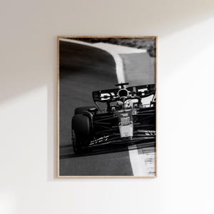 May include: Monochrome framed print of a Formula 1 car racing on a track. The car is captured mid-turn, with the track's white lines and the car's details in focus. The frame is a light wood.