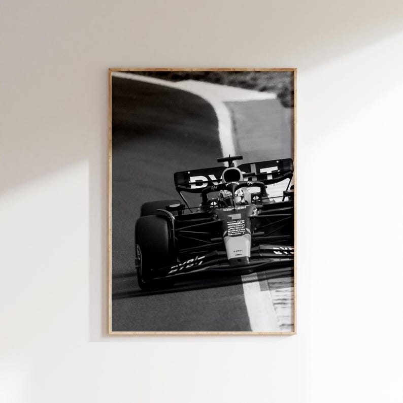 May include: Monochrome framed print of a Formula 1 car racing on a track. The car is captured mid-turn, with the track's white lines and the car's details in focus. The frame is a light wood.