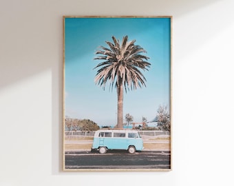 May include: A framed print featuring a light blue vintage van parked on a road beneath a tall palm tree. The sky is a bright blue, and the scene evokes a coastal or tropical vibe. The print is in a wooden frame.